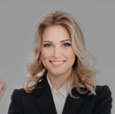 Smiling blonde woman in a black blazer and white blouse against a light gray background